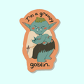 A sticker with an orange background sitting on op of a light blue background. The sticker is an illustration of a teal goblin sitting with their arms crossed and frowning. the sticker says "I'm a grumpy goblin." 